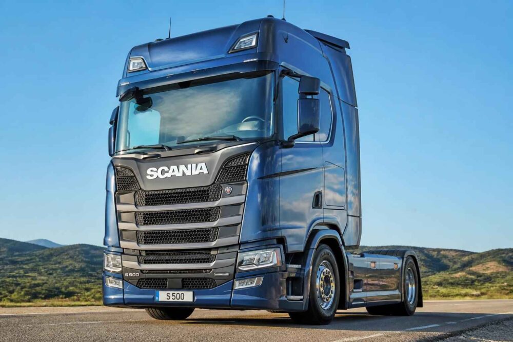 Niebieski ciągnik siodłowy Scania S 500 stojący na asfaltowej drodze wśród zielonych wzgórz