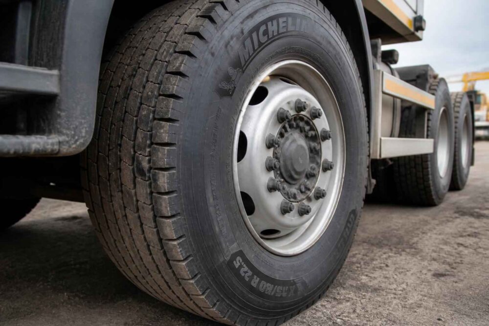 Zbliżenie na przednie koło ciężarówki z oponą Michelin i metalową felgą, w tle widoczne tylne osie pojazdu